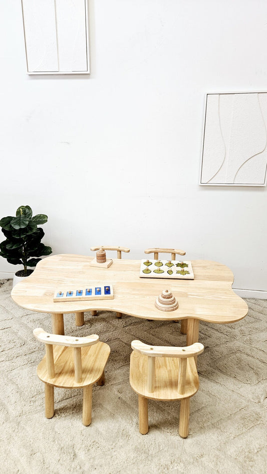 Wooden children's table and chairs set in a minimalistic room with white walls and a plant.